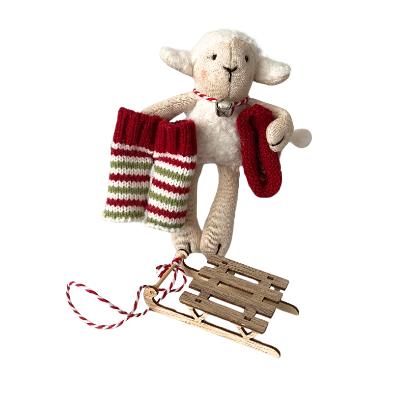 Fehér  szőrméjű kézműves textil bárány figura kezében kötött, csíkos nadrágja és kötött karácsonyi sapkája. Nyakában egy fém csengő, előtte egy fa szánkó.  
